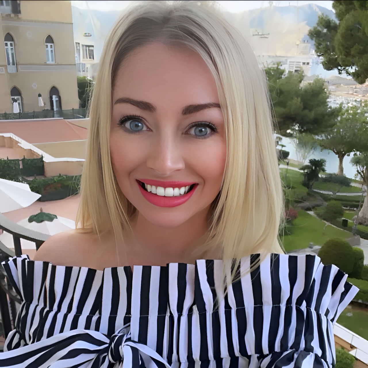 A cheerful blonde woman in an off-shoulder striped top with a scenic view behind her.