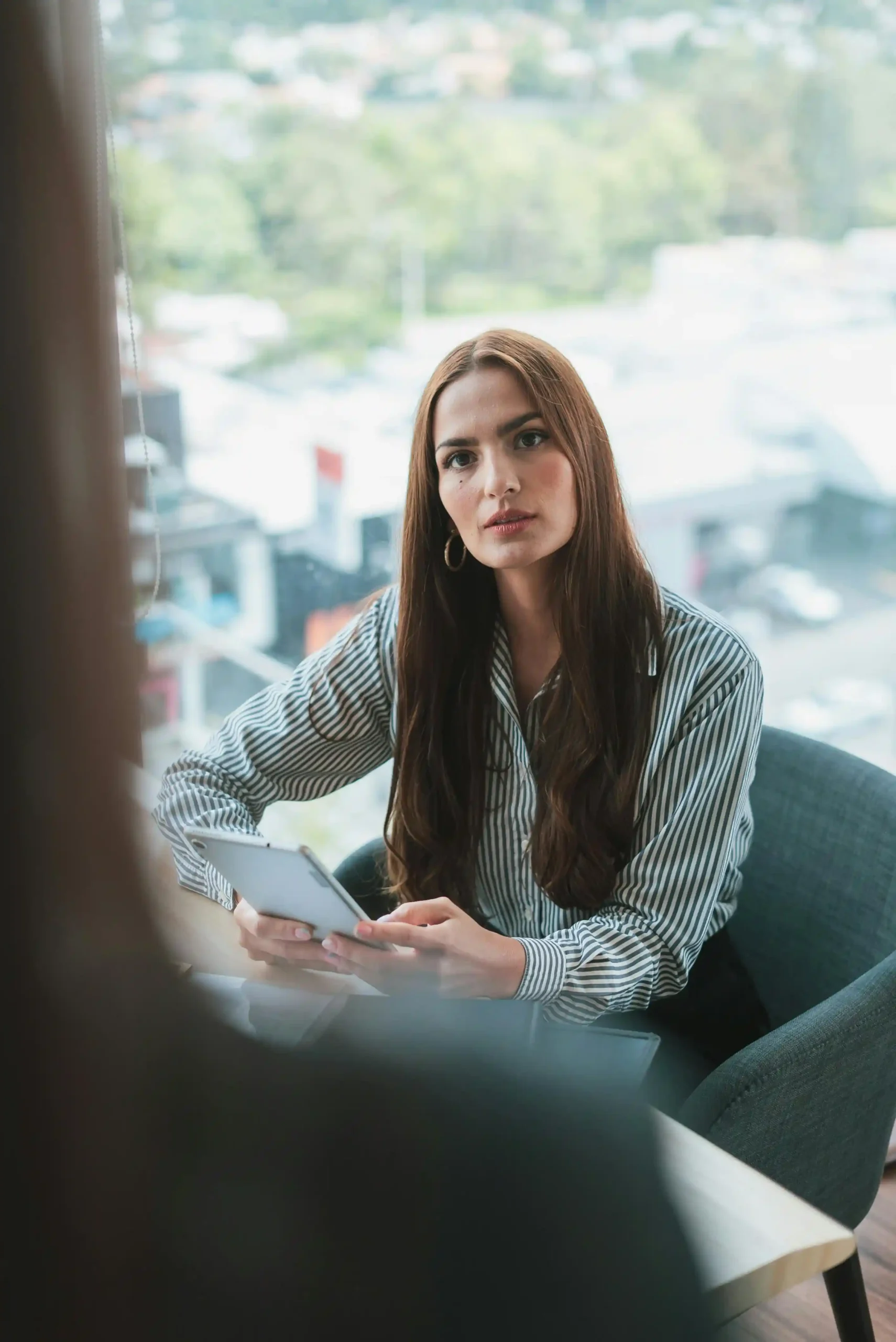 A woman in a striped shirt holding a tablet, sitting by a window during a conversation.