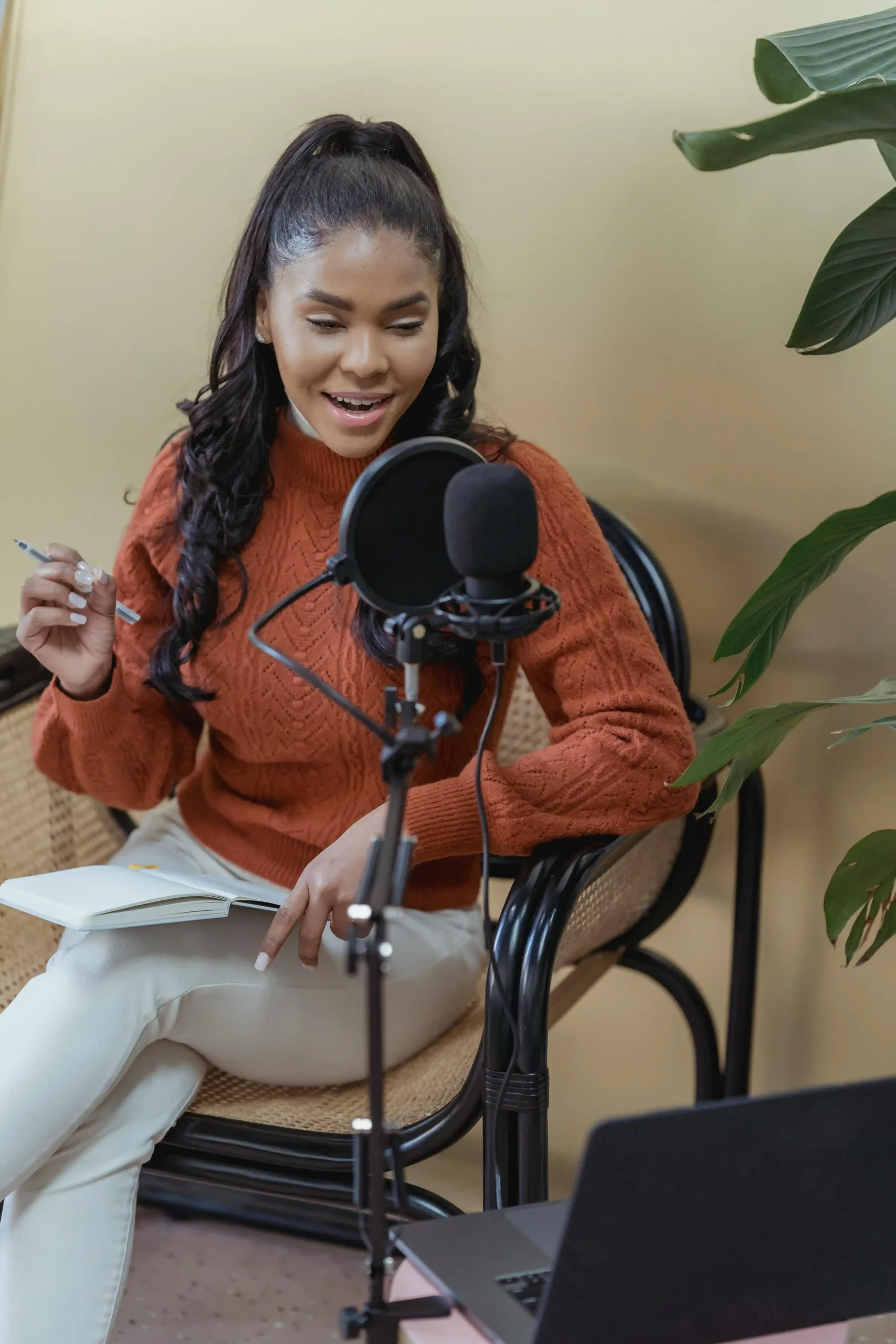 A woman in a rust-colored sweater speaking into a podcast mic while holding a notebook.