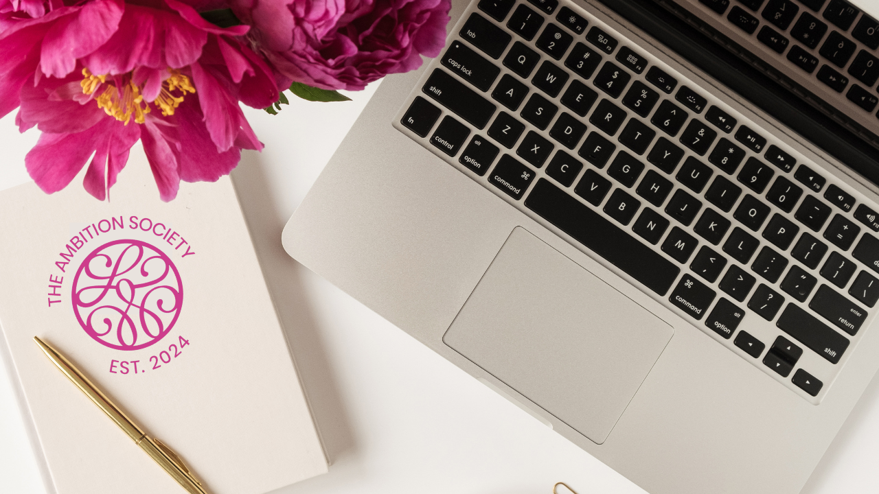 Flat lay of a laptop, notebook with "The Ambition Society Est. 2024" logo, and vibrant pink flowers.